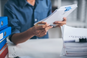 Person reviewing documents on a clipboard at a desk with stacked binders and paperwork, suggesting administrative or reporting work.