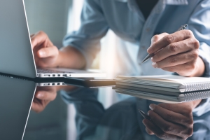 Person using a laptop while holding a pen over a notebook, with a reflective surface in the foreground.