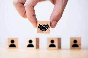 A hand placing a wooden block with a handshake icon at the front of a row of wooden blocks featuring human silhouette icons, symbolizing partnership or leadership in a team.