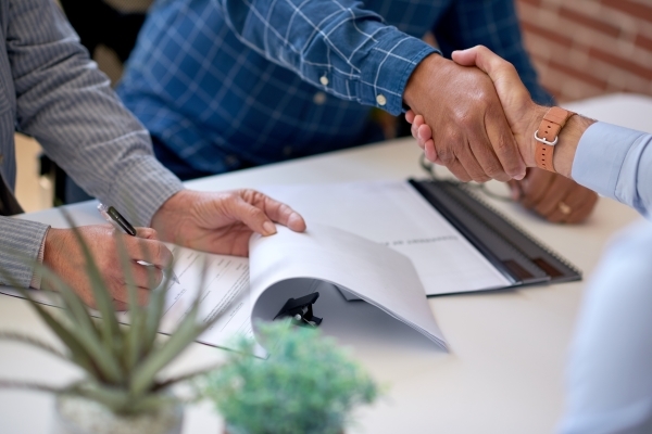Two people shake hands across a desk while another person signs paperwork, suggesting a business agreement or partnership.